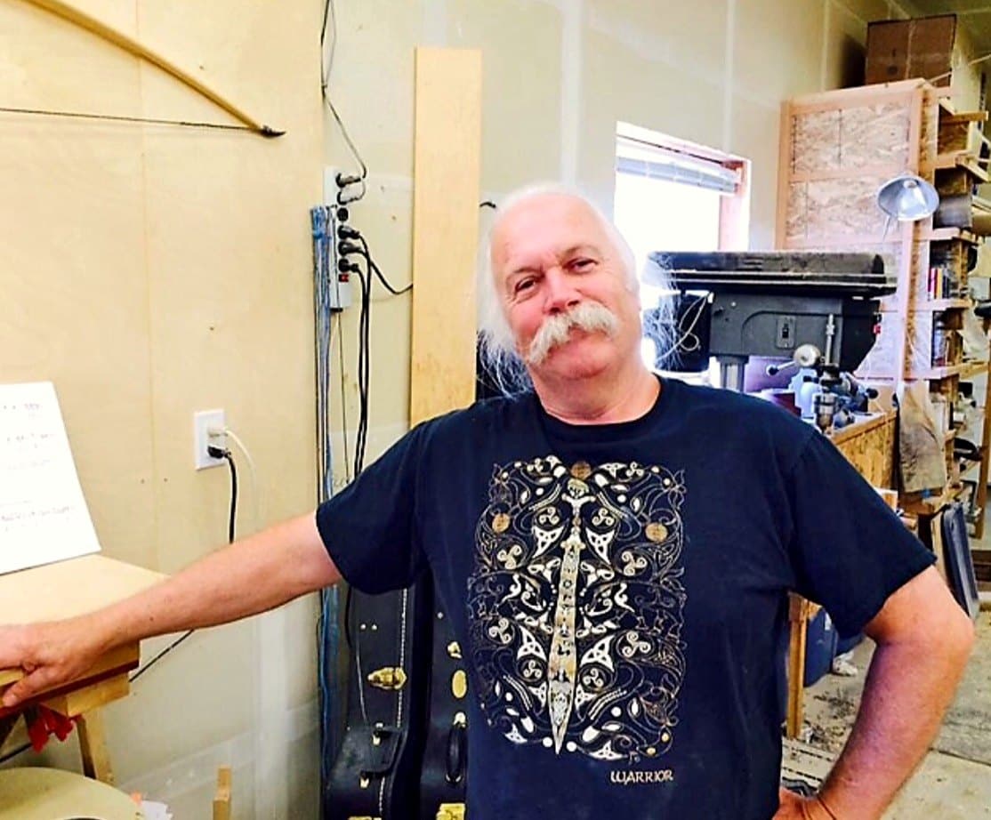 Doug Parry leaning on his workbench in his workshop, a longbow on the wall behind him